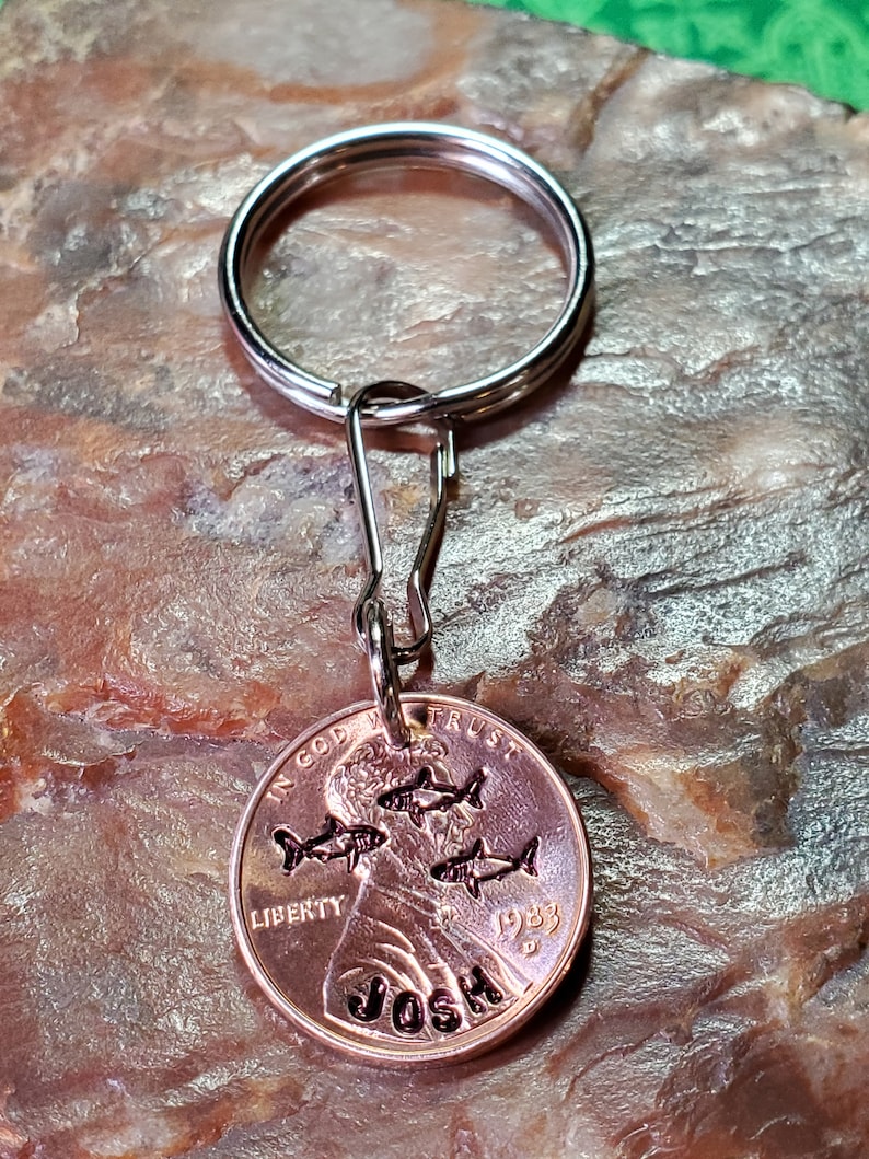 Op de afbeelding: Een koperen sleutelhanger met een zilveren sleutelring. De penny is gegraveerd met het woord "JOSH" en een ontwerp van vijf vissen. De penny is gedateerd 1983 en heeft de woorden "IN GOD WE TRUST" en "LIBERTY" erop gegraveerd.