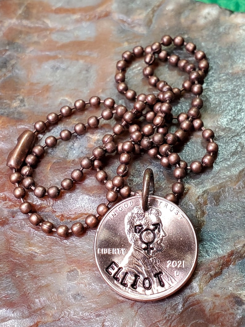 Puede incluir: Un collar de un centavo de cobre con una cadena de bolas. El centavo se ha personalizado con el nombre "Elliot" y un s&iacute;mbolo transg&eacute;nero. El a&ntilde;o 2021 tambi&eacute;n est&aacute; grabado en el centavo.