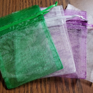 May include: Four sheer organza gift bags in shades of green, purple, and white, with drawstring closures. The bags are arranged on a wooden surface, showcasing their translucent texture and vibrant colors. Ideal for small gifts or favors.