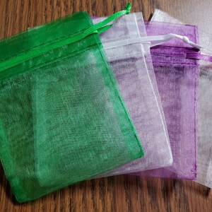 Op de afbeelding: Vier kleine organza zakjes in de kleuren groen, wit, paars en transparant. De zakjes zijn gemaakt van doorschijnend materiaal en hebben een trekkoord sluiting.