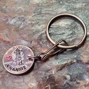 May include: A metal keychain featuring a coin with a lighthouse design, a red heart, and the name "FRANNIE." The coin is attached to a metal ring and clasp. The text "IN GOD WE TRUST" and "LIBERTY" are also visible.
