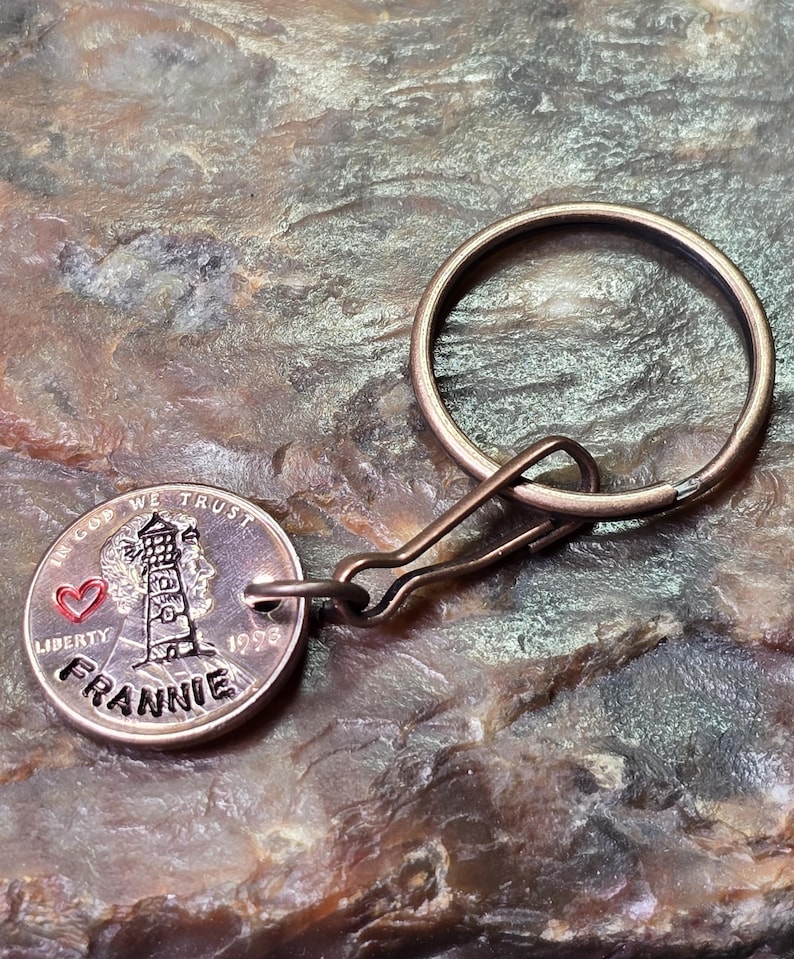 May include: A metal keychain featuring a coin with a lighthouse design, a red heart, and the name "FRANNIE." The coin is attached to a metal ring and clasp. The text "IN GOD WE TRUST" and "LIBERTY" are also visible.