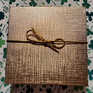 Puede incluir: Una caja de regalo dorada con una cinta dorada atada en un lazo.