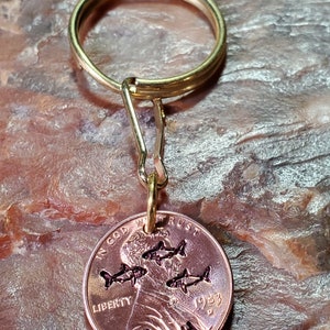 Op de afbeelding: Een gouden sleutelhanger met een 1983 penny eraan bevestigd. De penny heeft een ontwerp met vier vissen en de naam "Josh" erop gegraveerd.