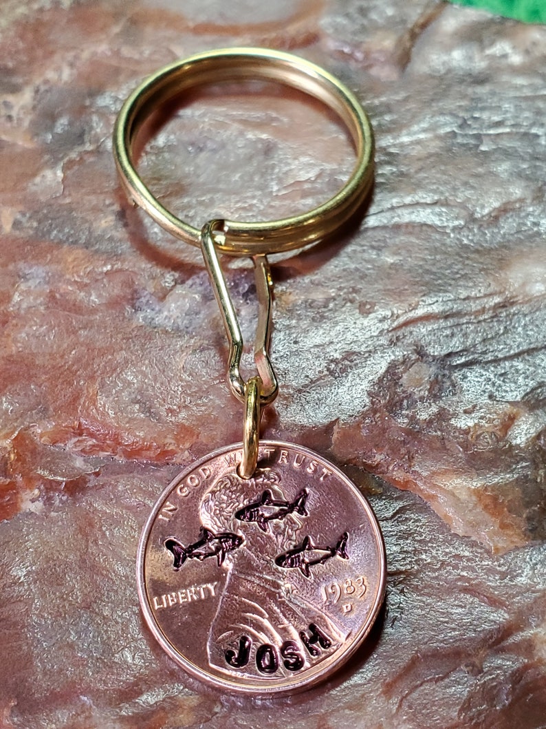 Op de afbeelding: Een gouden sleutelhanger met een 1983 penny eraan bevestigd. De penny heeft een ontwerp met vier vissen en de naam "Josh" erop gegraveerd.