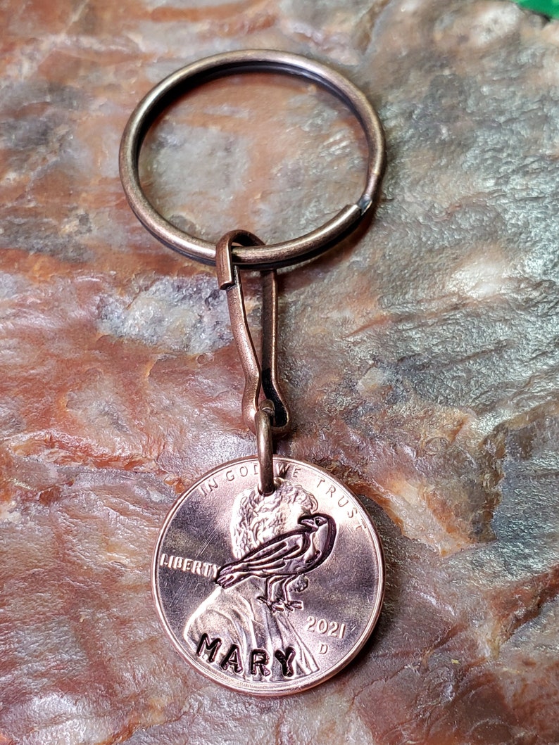 May include: A copper penny keychain with a personalized inscription of "MARY" and the year "2021". The penny features a design of a bird with the words "IN GOD WE TRUST" and "LIBERTY" on the coin.