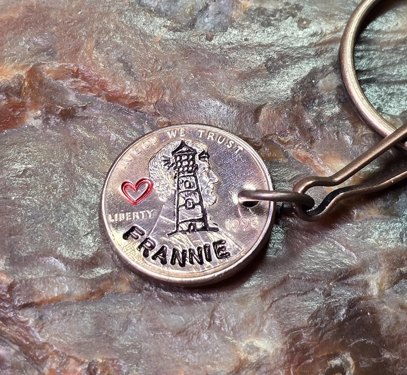 May include: A silver coin keychain featuring a lighthouse design with the name "FRANNIE" engraved below. The coin also has a red heart and the words "LIBERTY" and "IN GOD WE TRUST".
