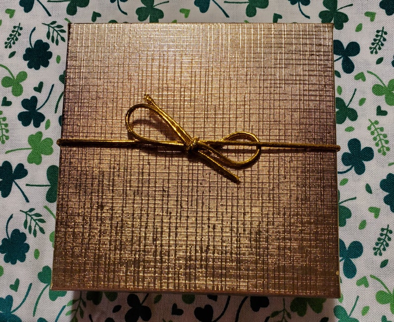 May include: A square, gold-colored gift box with a textured surface and a gold-colored bow tied around it. The box is sitting on a white fabric with green shamrocks.