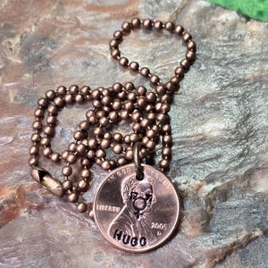Puede incluir: Un collar de cobre con una cadena de bolas. El centavo se ha personalizado con el nombre "HUGO" grabado en la parte posterior. El centavo est&aacute; fechado en 2005.