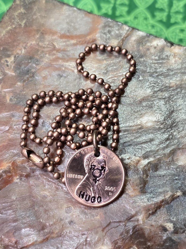 Puede incluir: Un collar de cobre con una cadena de bolas. El centavo se ha personalizado con el nombre "HUGO" grabado en la parte posterior. El centavo est&aacute; fechado en 2005.