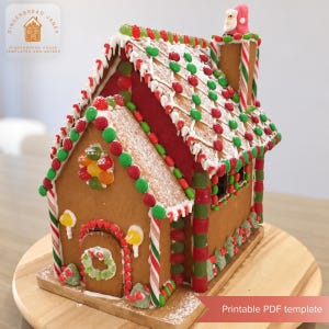 Op de afbeelding: Een peperkoekhuisje versierd met rode en groene snoepjes, wit glazuur en een kerstmanfiguur op de schoorsteen. Het dak heeft rijen kleurrijke lekkernijen en de voordeur heeft een kleine krans. Tekst leest Gingerbread Janey.