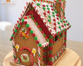 May include: A gingerbread house decorated with red and green candies, white icing, and a Santa figure on the chimney. The roof features rows of colorful sweets, and the front door has a small wreath. Text reads Gingerbread Janey.