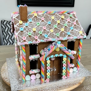 Op de afbeelding: Een gingerbreadhuis versierd met kleurrijke snoepjes en sprinkles. Het huis heeft een wit glazuur dak met een snoepstok ontwerp en een schoorsteen. De muren zijn versierd met kleurrijke snoepjes en sprinkles. Het huis heeft een deur en ramen.