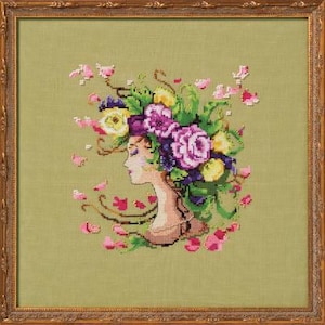 May include: A cross-stitch embroidery of a woman's profile with a floral crown of purple, yellow, and pink flowers. The background is a light green fabric.