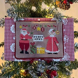 May include: A framed cross-stitch Christmas ornament featuring Santa and Mrs. Claus. The ornament has a red and white gingham border with lace trim. The cross-stitch design includes the text "MERRY XMAS" and festive decorations.