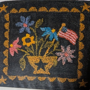 May include: A black rug with a yellow border and a design of a vase with flowers and an American flag. The vase is brown and has a star in the center. The flowers are blue, pink, yellow, and white.