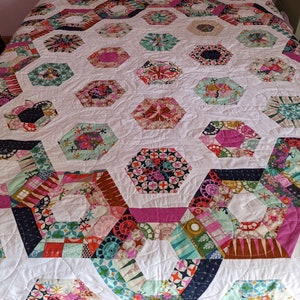 May include: A handmade quilt with a white background and colorful, hexagonal patchwork designs. The quilt features various patterns and colors, including pink, blue, green, and floral prints. The quilt is on a bed, showcasing its size and design.