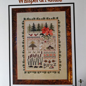 May include: Cross-stitch pattern titled "Whisper of Nature" featuring a detailed nature scene. The design includes trees, flowers, birds, bees, and the word "Buzz." The artwork is framed with a brown border and a decorative green border.
