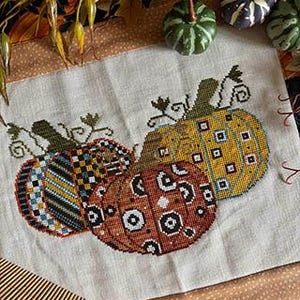May include: A cross-stitch embroidery on light beige fabric featuring three pumpkins with distinct patterns. One pumpkin is orange with black and white circles, one is yellow with blue squares, and one has black, white, and orange stripes.