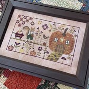 May include: A framed cross-stitch sampler featuring a fall scene with a pumpkin house, squirrel, and bird. The artwork is in a dark brown frame and rests on a colorful patchwork quilt. The sampler has a light pink background with various fall-themed elements.