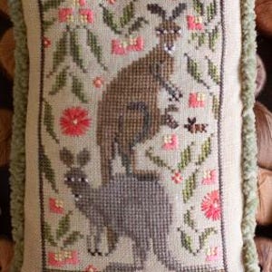May include: A beige cushion with a green fringed border features an embroidered design of two kangaroos in a floral setting. The larger kangaroo is brown and the smaller kangaroo is grey. The floral design includes pink, green and white flowers.