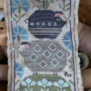 May include: A hand-stitched pillow featuring a whimsical design of three stacked pigs with a bird perched on top. The pigs are embroidered in shades of brown, gray, and white, with a green and blue floral pattern surrounding them.