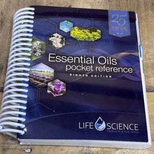 May include: A spiral-bound book titled "Essential Oils pocket reference, Eighth Edition." The cover is dark blue with images of flowers and plants. The book also features the text "25 Years Young" and the Life Science Products & Publishing logo.
