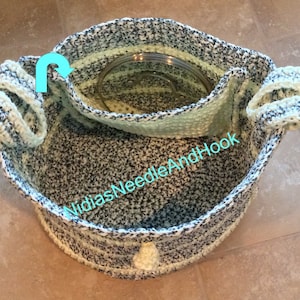 以下が含まれることがあります： 黒と白の斑点模様の糸と明るい緑色のストライプで編まれた、かぎ針編みのトートバッグ。バッグには2つのハンドルと、底に小さなかぎ針編みのポンポンが付いています。バッグは裏返して内側を見せるようにしています。バッグの内側には「Nidi's Needle And Hook」という文字が見えます。