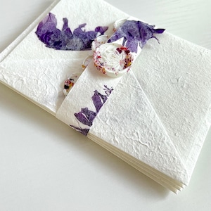 May include: A stack of white envelopes with a textured surface. The envelopes are decorated with pressed purple flowers and a white wax seal with a floral design.
