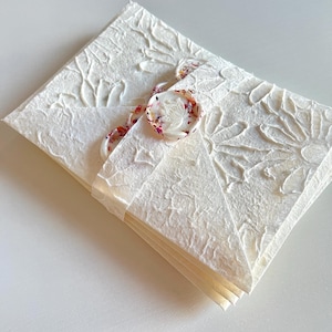 May include: A white envelope with a floral embossed design and a white wax seal with a floral design. The envelope is tied with a ribbon with a floral pattern.