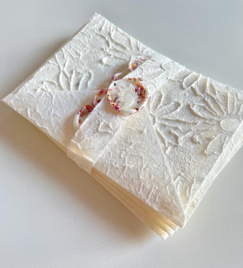 May include: A white envelope with a floral embossed design and a white wax seal with a floral design. The envelope is tied with a ribbon with a floral pattern.