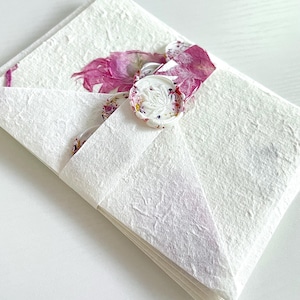 May include: A stack of white envelopes with a textured surface. The envelopes are tied together with a white ribbon and have a white wax seal with a floral design. The seal is decorated with dried flowers.