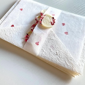 May include: A stack of white paper envelopes with a red heart design. The envelopes are tied together with a white ribbon and a wax seal with a heart design.