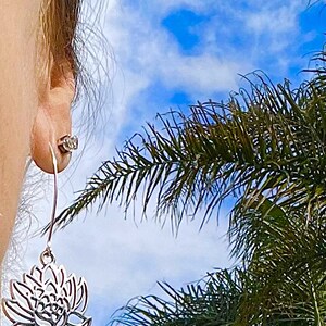 May include: Silver dangle earrings with a delicate lotus flower design. The earrings are shown against a blue sky with palm trees in the background.
