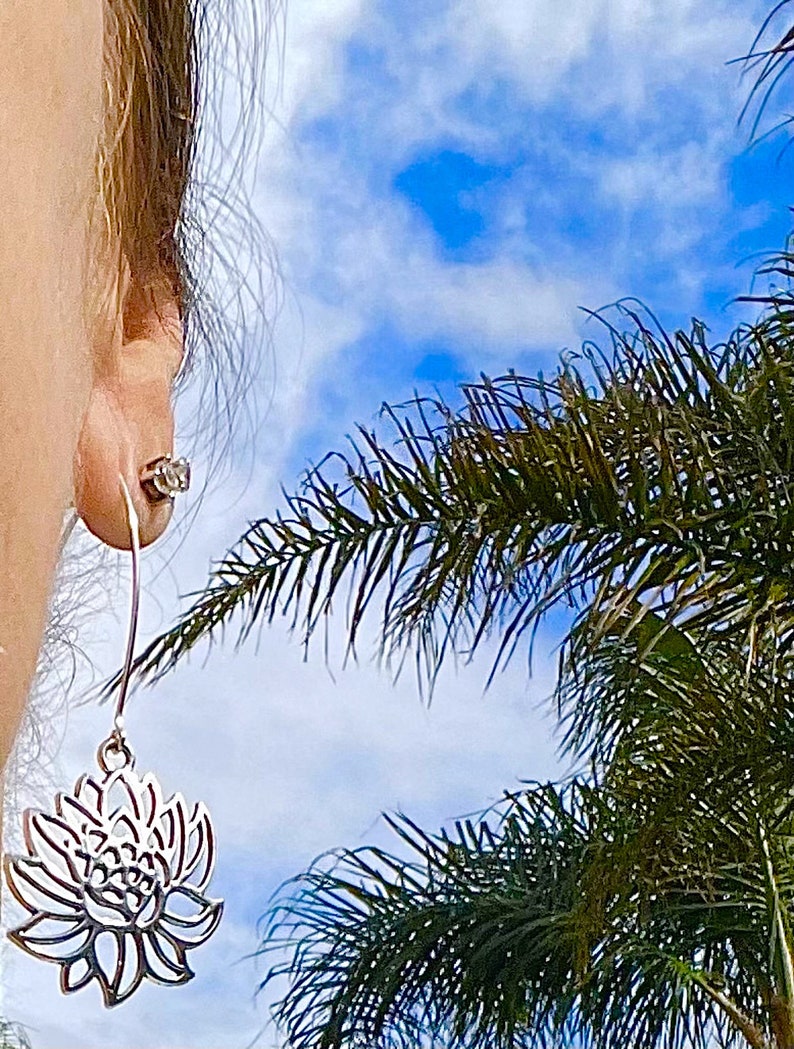 May include: Silver dangle earrings with a delicate lotus flower design. The earrings are shown against a blue sky with palm trees in the background.