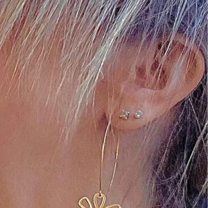 May include: Gold-colored flower-shaped dangle earrings. The earrings are hanging from a person's ear. The earrings are a floral design. The image also contains the text: "Also available in Gold! See listing on my Shop."