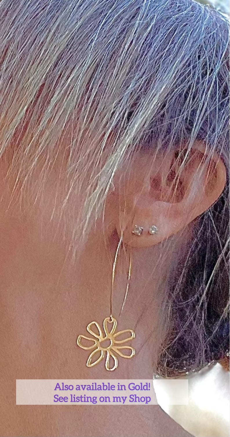 May include: Gold-colored flower-shaped dangle earrings. The earrings are hanging from a person's ear. The earrings are a floral design. The image also contains the text: "Also available in Gold! See listing on my Shop."