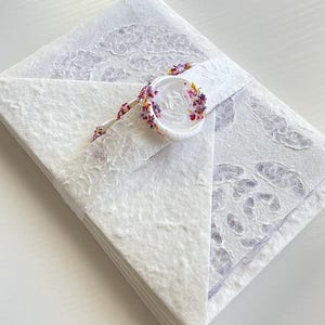 May include: A stack of white handmade paper envelopes, sealed with a white wax seal adorned with dried flower petals. The envelopes are wrapped with a matching paper band.
