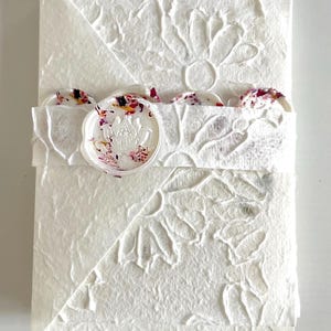 May include: White envelope with a decorative floral pattern and a wax seal with a floral design. The envelope is made of textured paper.