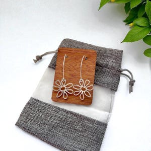 May include: Silver daisy earrings displayed on a wooden plaque inside a gray drawstring bag. The earrings have a delicate, openwork design. The bag is made of a textured gray fabric with a white lining. The background is a soft white.