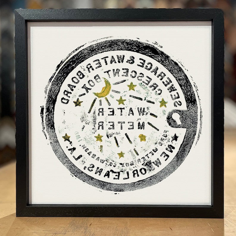 Puede incluir: Imagen en blanco y negro de una tapa de alcantarilla circular con el texto "NEW ORLEANS SEWERAGE & WATER BOARD CRESCENT BOX" y una luna creciente con estrellas.