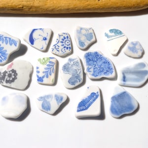 Puede incluir: Un conjunto de fragmentos cerámicos de forma irregular, predominantemente blancos con diseños florales y abstractos azules. Los fragmentos varían en tamaño y diseño, con un palo de madera en la parte superior. Dispuestos sobre una superficie blanca.