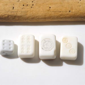 May include: A collection of ivory-colored game pieces, including a die and three rectangular tiles. The tiles feature etched designs, such as sets of symbols and a circular pattern. A piece of driftwood rests above the game pieces.