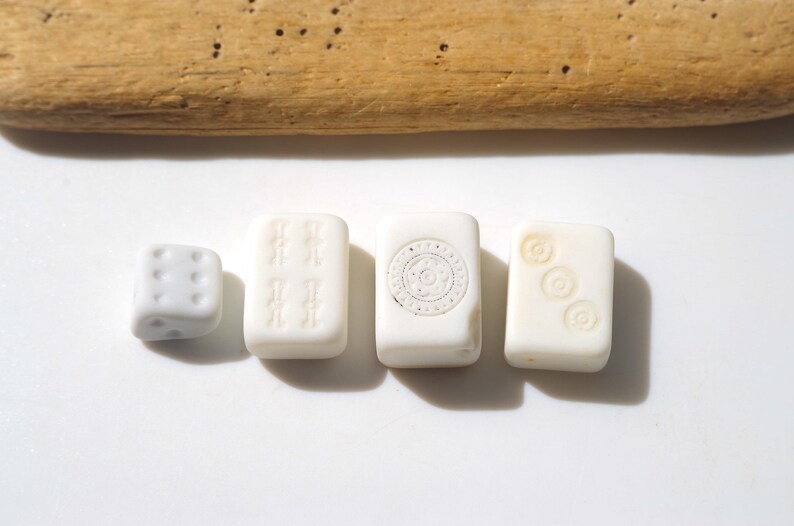 May include: A collection of ivory-colored game pieces, including a die and three rectangular tiles. The tiles feature etched designs, such as sets of symbols and a circular pattern. A piece of driftwood rests above the game pieces.