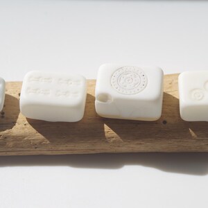 May include: Four white, square-shaped objects with different embossed designs are arranged on a piece of driftwood. The objects include a die with six dots, a block with text, a block with a circular design, and a block with three circles.