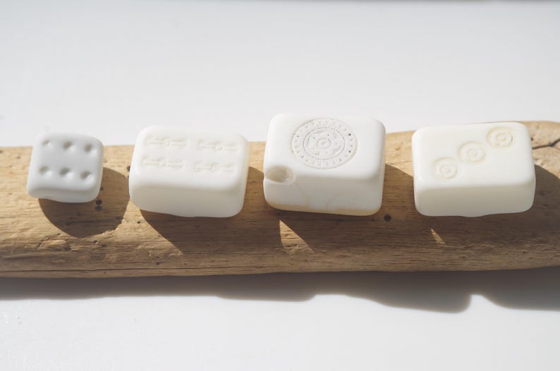 May include: Four white, square-shaped objects with different embossed designs are arranged on a piece of driftwood. The objects include a die with six dots, a block with text, a block with a circular design, and a block with three circles.