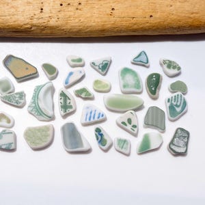 May include: A collection of small, smooth sea glass shards in various shades of green, white, and blue, featuring assorted patterns and textures. The pieces are arranged in a loose rectangular formation below a single piece of weathered driftwood.