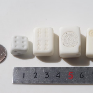 May include: A penny, a white die, and three white rectangular game tiles are arranged in a row. The tiles have carved designs. A metal ruler is placed below the objects, showing measurements in inches.