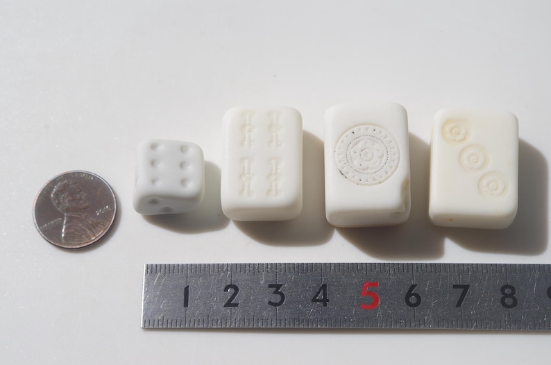 May include: A penny, a white die, and three white rectangular game tiles are arranged in a row. The tiles have carved designs. A metal ruler is placed below the objects, showing measurements in inches.