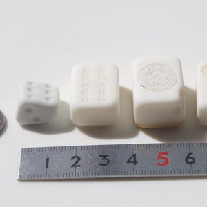 May include: A collection of small, white game pieces, including a die and several rectangular tiles, are arranged on a white surface. A ruler shows measurements in inches. A penny is also included for size comparison.